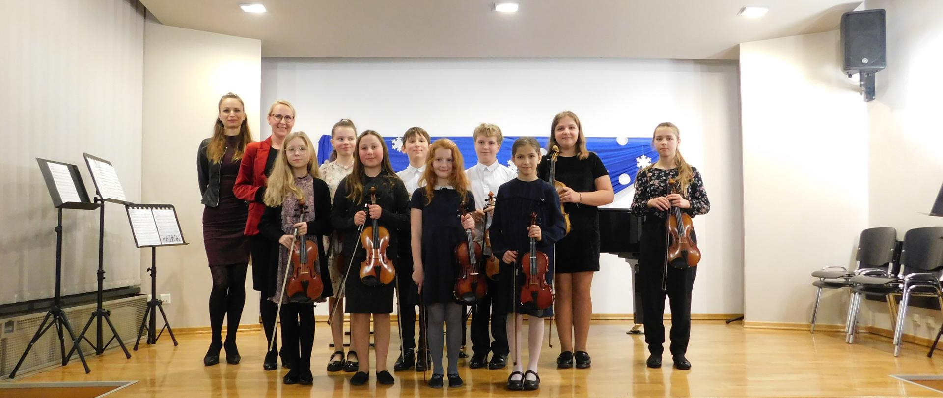 Na scenie młodzi artyści koncert klasowego. Uczniowie wraz z nauczyciele i akompaniatorem. Wszyscy uczniowie trzymają w rękach skrzypce.