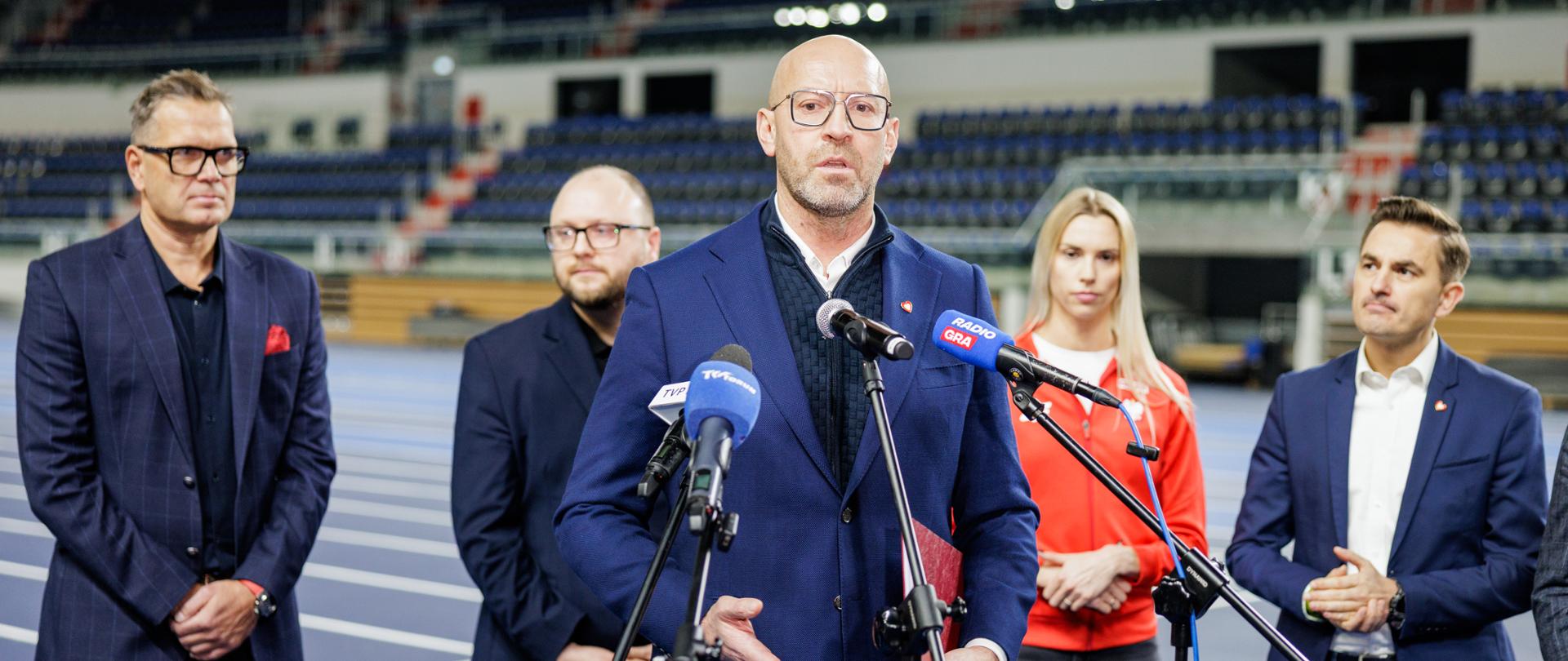 Konferencja prasowa na bieżni Areny Toruń. Minister sportu i turystyki Jakub Rutnicki mówi do mikrofonu. Za nim stoją inni uczestnicy konferencji. W tle widać trybuny.