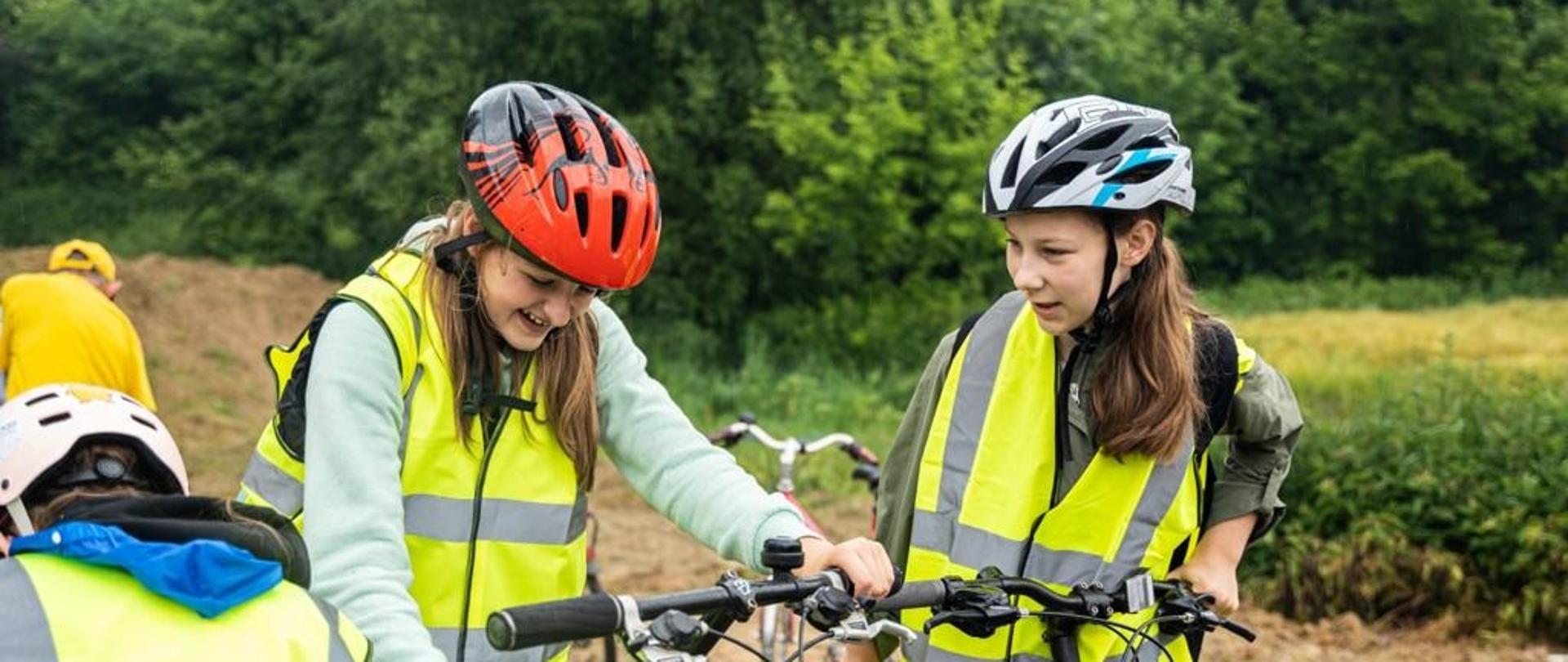 Pada deszcz, na wyłożonej szarą kostką drodze kilkoro dzieci w żółtych kamizelkach siada na rowery, za nimi zielone drzewa.