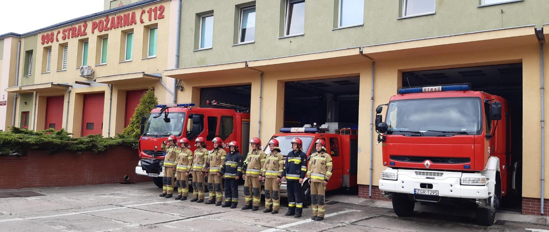 Strażacy stojący w szeregu. Z tyłu wozy strażackie na tle komendy.