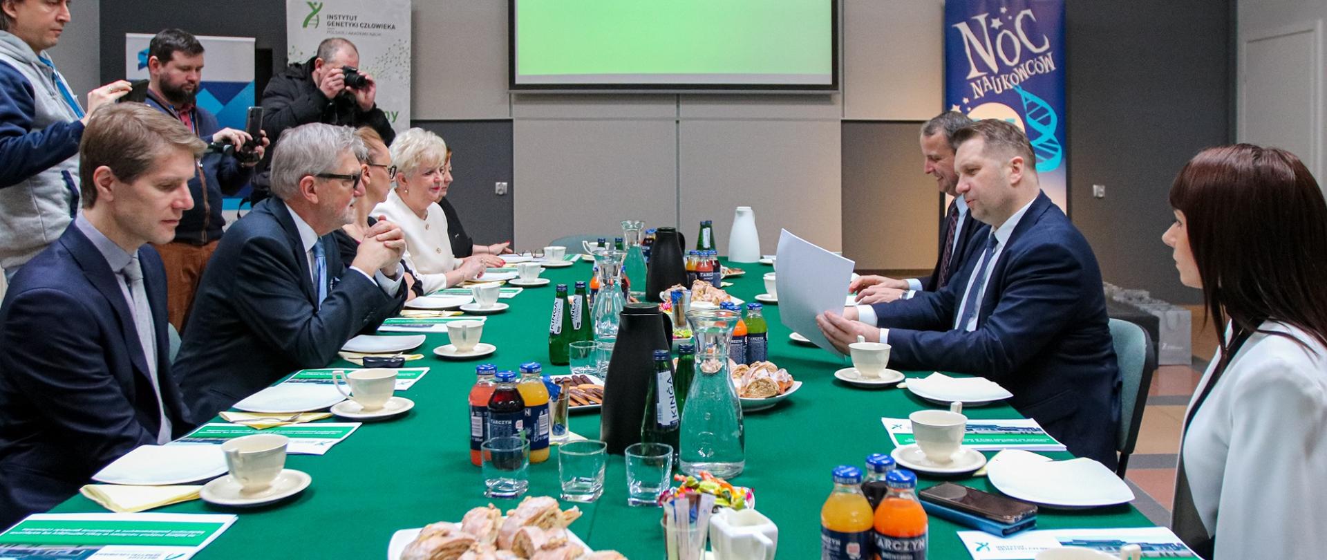 Po dwóch stronach długiego, nakrytego zielonym obrusem stołu siedzą elegancko ubrani ludzie, nad stołem na ścianie wisi duży monitor.