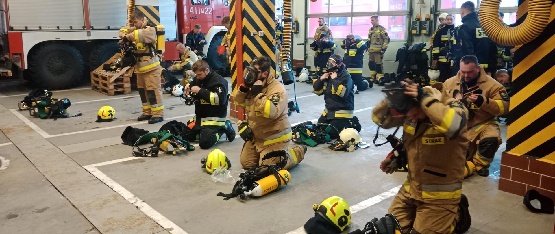 Kolorowa fotografia wykonana w pomieszczeniu w pogodny dzień w strażnicy. Przedstawia strażaków w ubraniach specjalnych w pozycji klęczącej ubierających sprzęt ochrony układu oddechowego.
