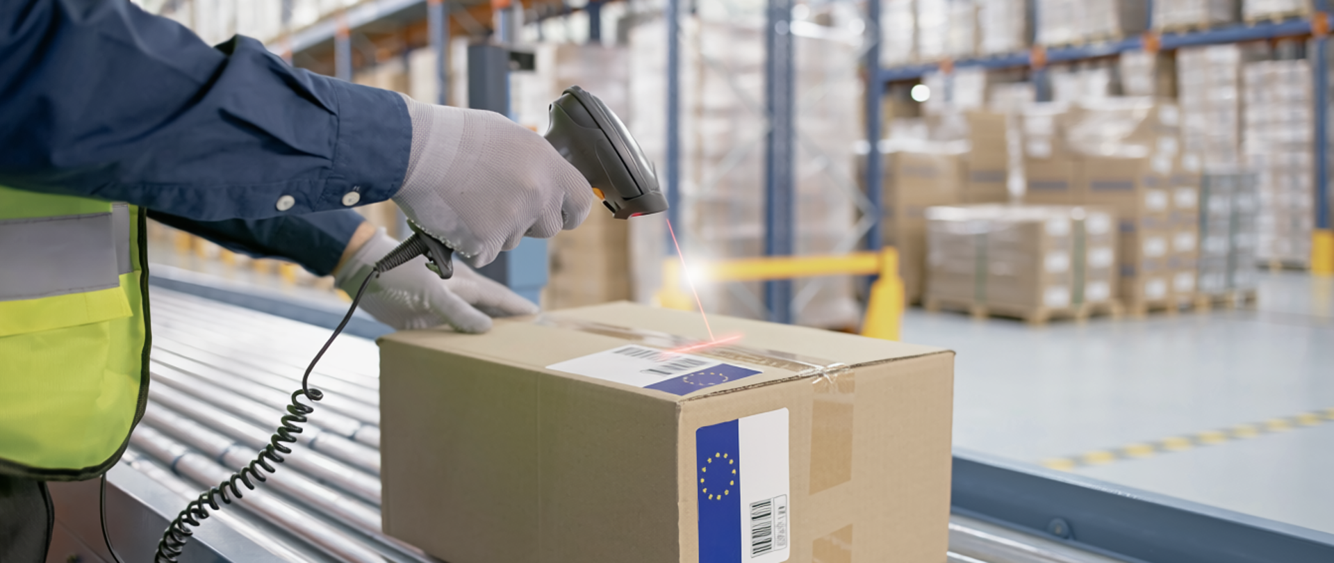 Osoba w kamizelce odblaskowej skanuje kod kreskowy na kartonie, leżącym na taśmie transportowej w magazynie pełnym regałów z zapakowanymi przesyłkami.
