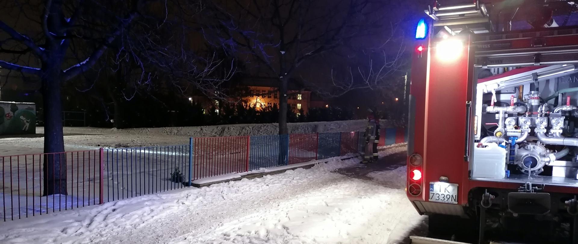 Zdjęcie przedstawia samochód strażacki który znajduje się przed ogrodzeniem posesji przedszkola. Po lewej stronie widać budynek przedszkola - nie widać ognia ani dymu