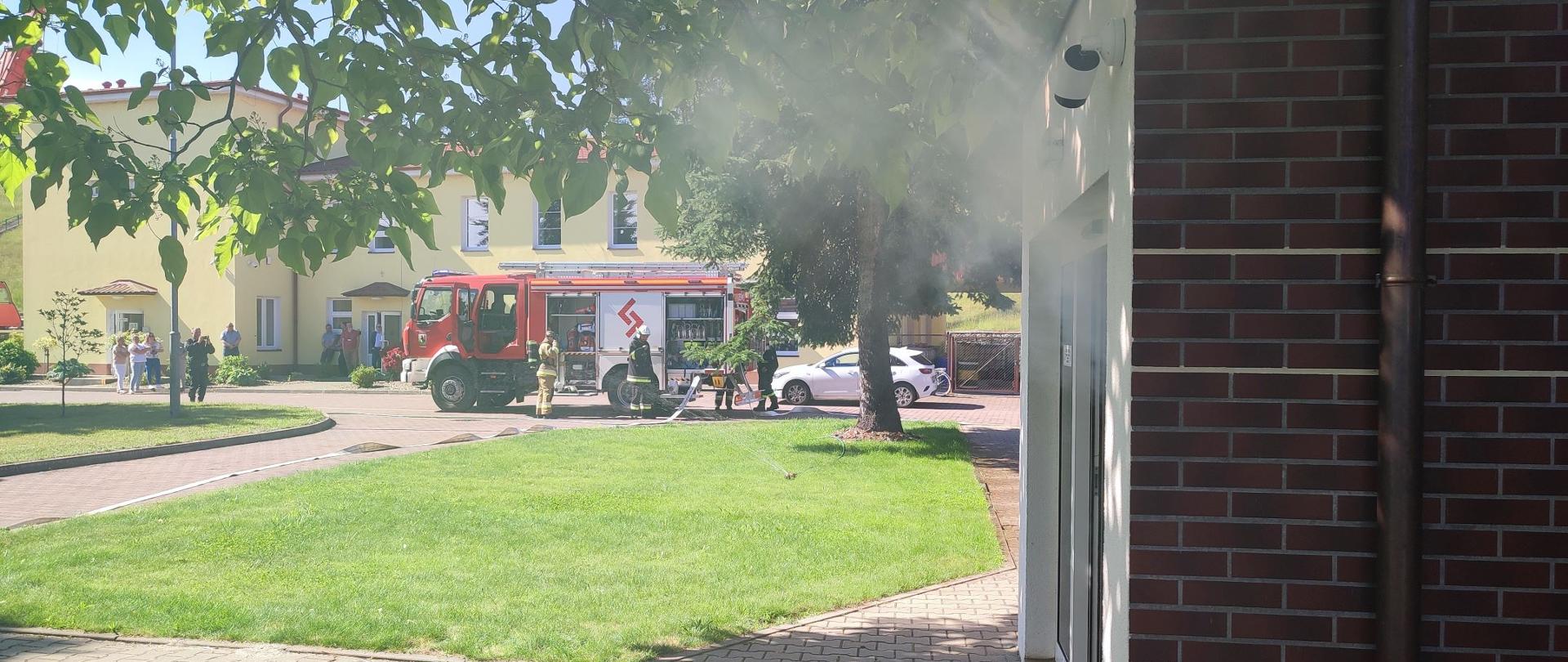 Na zdjęciu znajduje się samochód pożarniczy przy którym stoją strażacy. Pojazd znajduje się na terenie elektrowni wodnej Dychów. Widoczni także są pracownicy zakładu i dym wydobywający się z drzwi.
