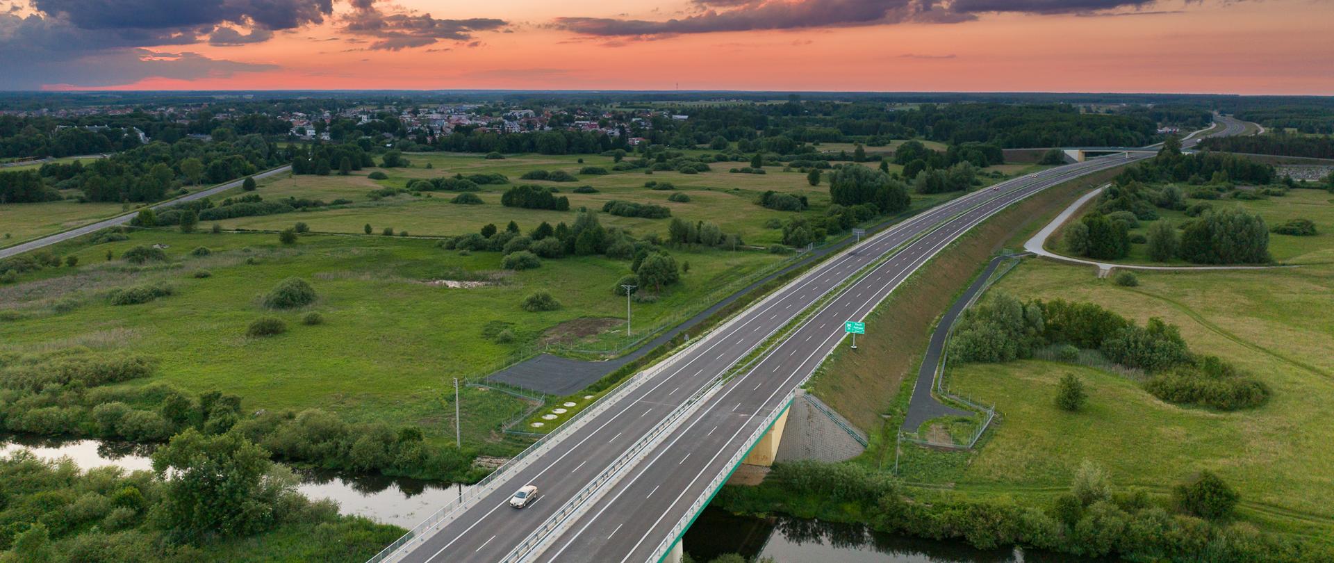 Widok z lotu ptaka na drogę ekspresową S19 przecinającą zielone pola i rzekę; w tle zabudowania i zachód słońca z kolorowymi chmurami.