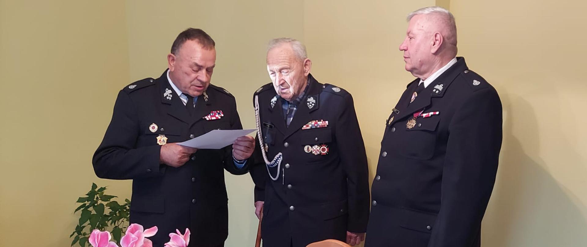 Zdjęcie przedstawia trzech druhów OSP w mundurach galowych. Druh Tadeusz Niedźwiecki odczytuje list gratulacyjny z okazji 98 urodzin dla stojącego obok druha Franciszka Flaka.