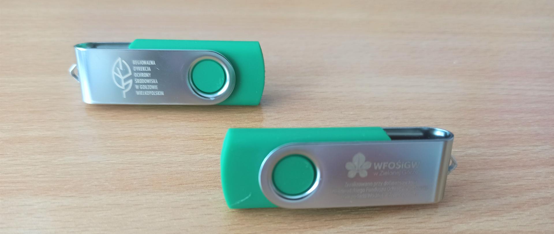 Na brązowym blacie leżą dwa zielono-srebrne pendrive 'y. Na pendrive'ach umieszczono napisy: Regionalna Dyrekcja Ochrony Środowiska w Gorzowie Wielkopolskim oraz Wojewódzki Fundusz Ochrony Środowiska i Gospodarki Wodnej w Zielonej Górze 