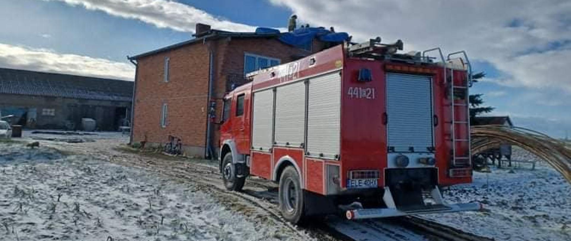 Na zdjęciu widać stojący na polnej drodze czerwony samochód strażacki ustawiony przodem w stronę zabudowań. W tle widać budynek mieszkalny z czerwonej cegły. Na dachu, który uległ zniszczeniu, strażak w żółtym stroju i czerwonym hełmie zakłada niebieską plandekę. Dom zbudowany jest z czerwonej cegły. Dalej w tle widać murowany budynek gospodarczy. Obok samochodu strażackiego znajdują się pola. Po prawej stronie widać zebrane konstrukcje metalowe od tunelu rolniczego. Jest widno.