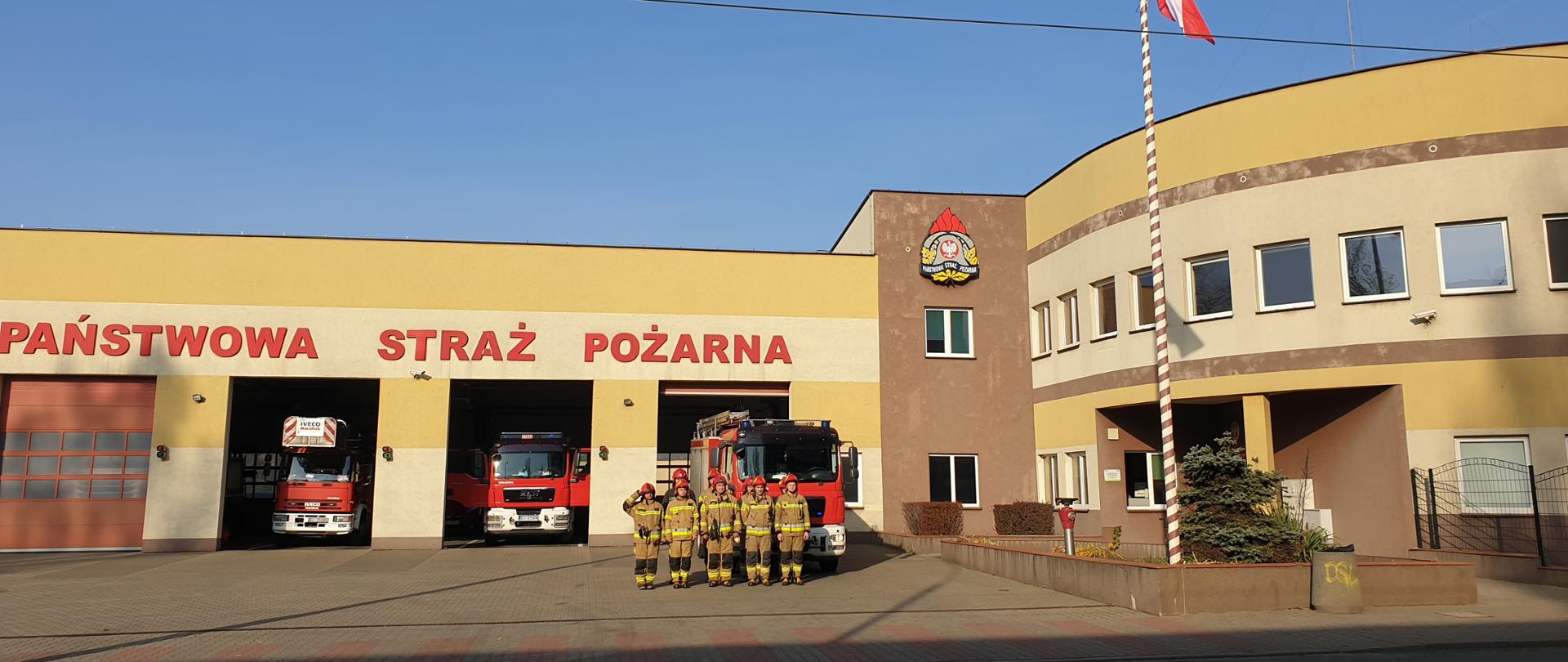 Strażacy stojący na baczność obok masztu z podniesioną flagą biało-czerwoną, w tle jednostka ratowniczo-gaśnicza oraz samochody bojowe