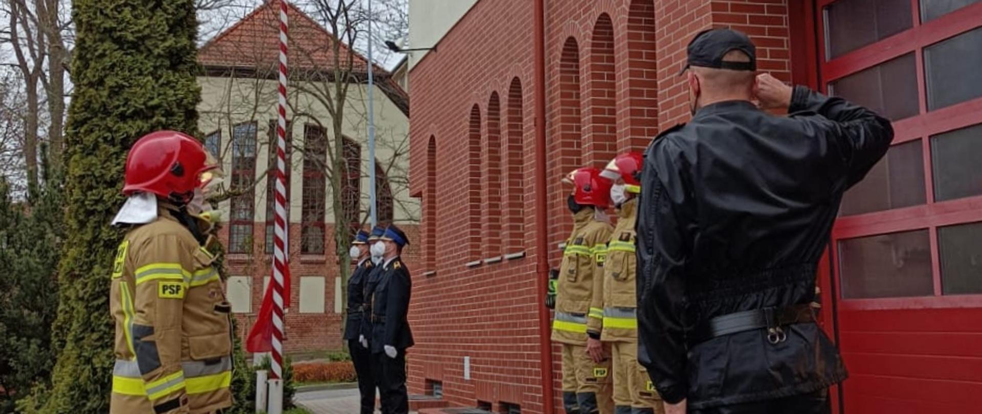 strażacy w mundurach specjalnych stoją w dwóch szeregach na przeciw siebie. Dalej poczet flagowy wciąga flagę na masz. Dowódca salutuje