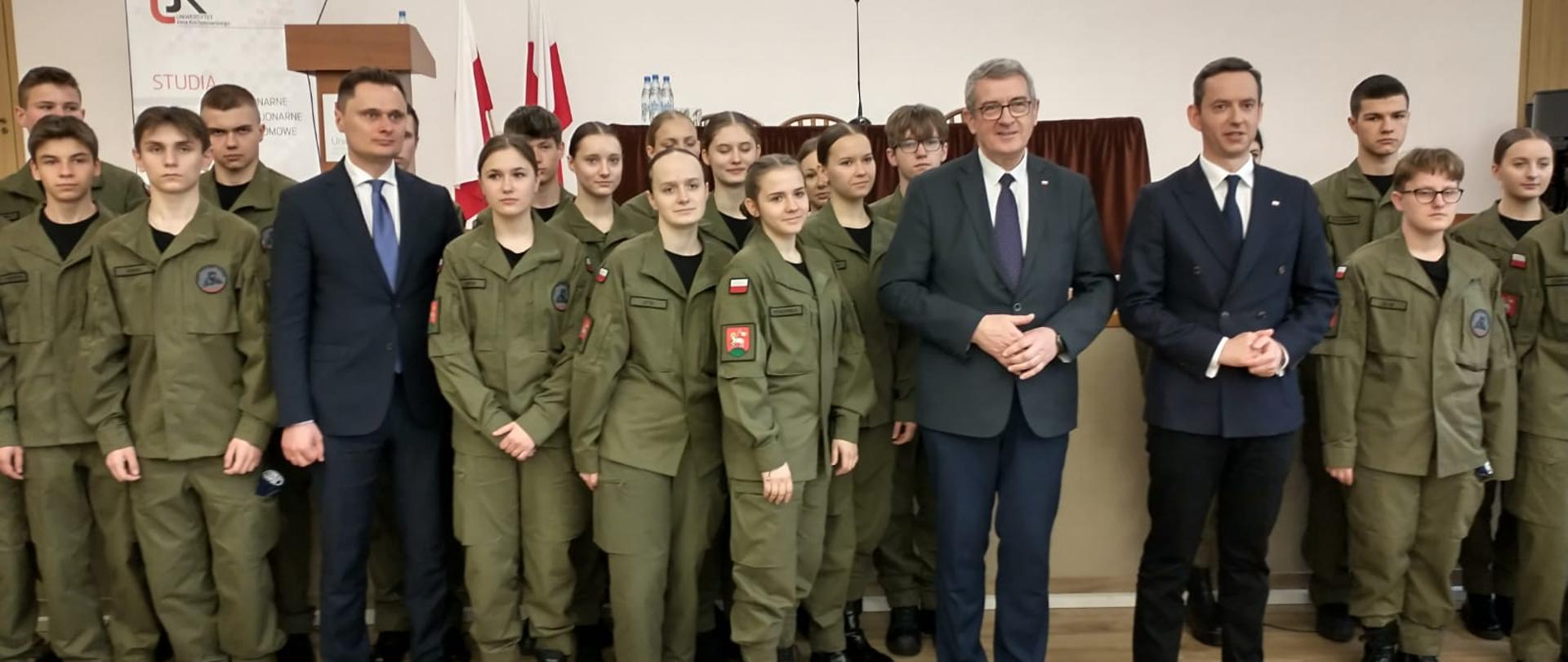 Na sali z drewnianą podłogą stoi duża grupa młodzieży w zielonych mundurach, między nimi wiceminister Murdzek i dwóch mężczyzn w garniturach.