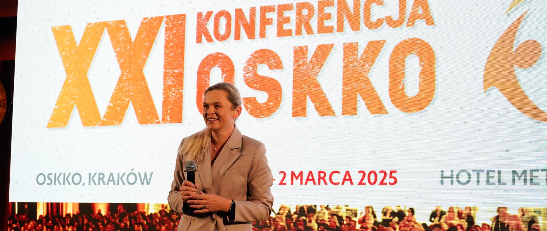 XXI Konferencja OSKKO, barbara Nowacka przemawia do mikrofonu na stojąco do publiczności. Za nią ekran z tytułem konferencji