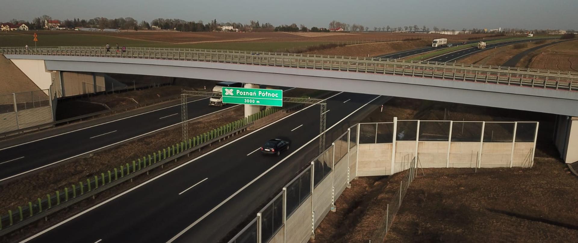 Zdjęcie przedstawia drogę ekspresową. Widok na węzeł Poznań Północ. Pomiędzy jezdniami widoczny jest pas rozdziału. Nad drogą widoczny jest wiadukt drogowy. W górnej lewej części zdjęcia widoczne zabudowania mieszkalne. Po prawej stronie widoczne elementy brd takie jak: ekrany akustyczne. 