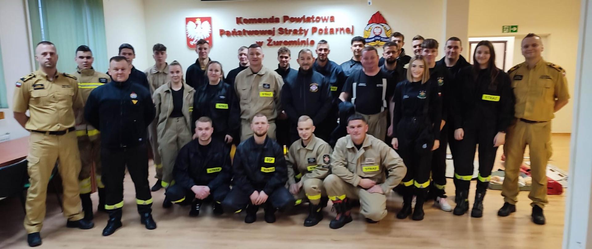 Zdjęcie przedstawia grupę strażaków oraz uczestników szkolenia pozujących do wspólnej fotografii w sali Komendy Powiatowej Państwowej Straży Pożarnej w Żurominie. Na pierwszym planie widoczny jest fantom medyczny, 