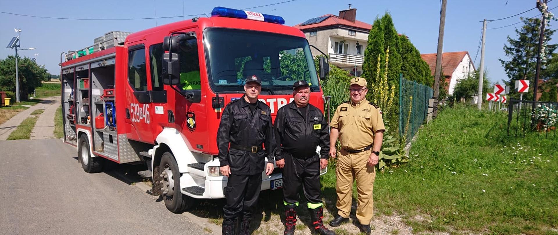 pora dzienna, trzech umundurowanych strażaków stoi przed samochodem pożarniczym