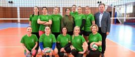 Zdjęcie przedstawia żeńską drużynę siatkarską pozującą do wspólnej fotografii w hali sportowej. W dolnym rzędzie 5 kobiet klęczy lub kuca. Trzy z nich trzymają piłki do siatkówki. Zawodniczki są uśmiechnięte, ubrane w zielone koszulki. W rzędzie górnym stoi 7 kobiet, ubranych w zielone koszulki, jedna z nich ma zielona bluzę. Po prawej stronie stoi mężczyzna w garniturze. Zdjęcie zrobione jest na tle siatki do siatkówki. W tle widać ściany hali wyłożone panelami oraz fragmenty trybun.