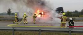 trwa gaszenie samochodu dostawczego marki Iveco stojącego na pasie awaryjnym autostrady, dwóch strażaków w rocie podaje prąd wody na całkowicie objęty płomieniami samochód, przed samochodem trzech strażaków asystujących, akcja odbywa się na całkowicie zablokowanym pasie autostrady