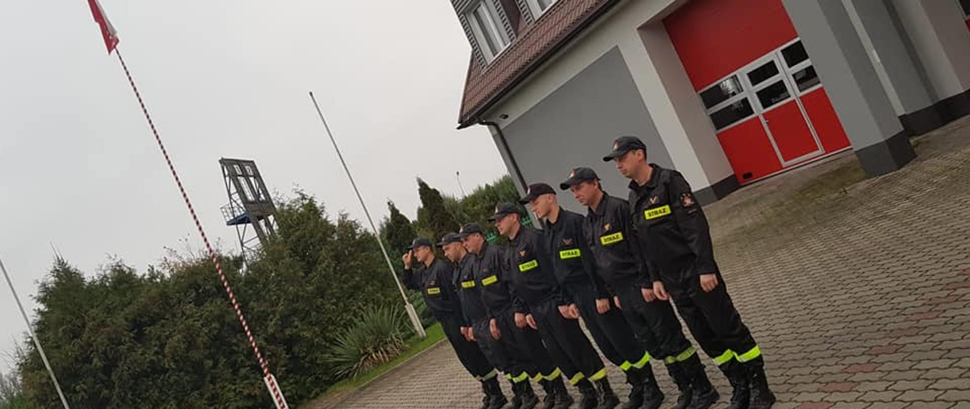 Na zdjęciu widać siedmiu strażaków ubranych w czarne stroje z jasnozielonymi odblaskami z napisem straż. Na głowie mają założone czapki z daszkiem. Mężczyźni stoją w jednym rzędzie. W tle widać biało-czerwony maszt na którym zawieszona jest biało-czerwona flaga, budynek komendy oraz wspinalnie. Zdjęcie jest pochylone. Jest widno.