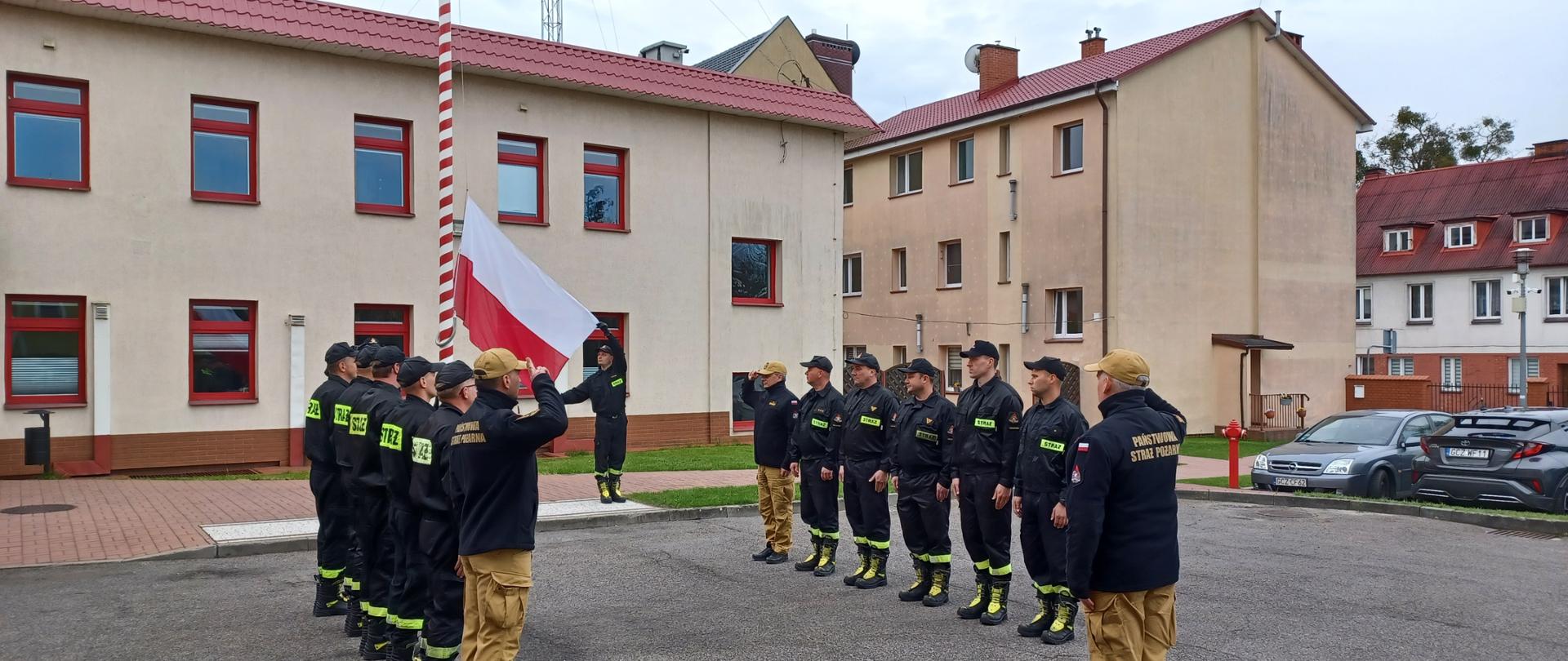 Uroczysta zmiana służby z okazji Dnia Flagi Rzeczpospolitej Polskiej. Zmiany służbowe stoją naprzeciw siebie. Na szczycie pododdziału stoi Komendant Powiatowy Państwowej Straży Pożarnej w Człuchowie st. bryg. Radosław Batruch. Poczet flagowy stoi przy maszcie, następuje uroczyste podniesienie flagi państwowej.