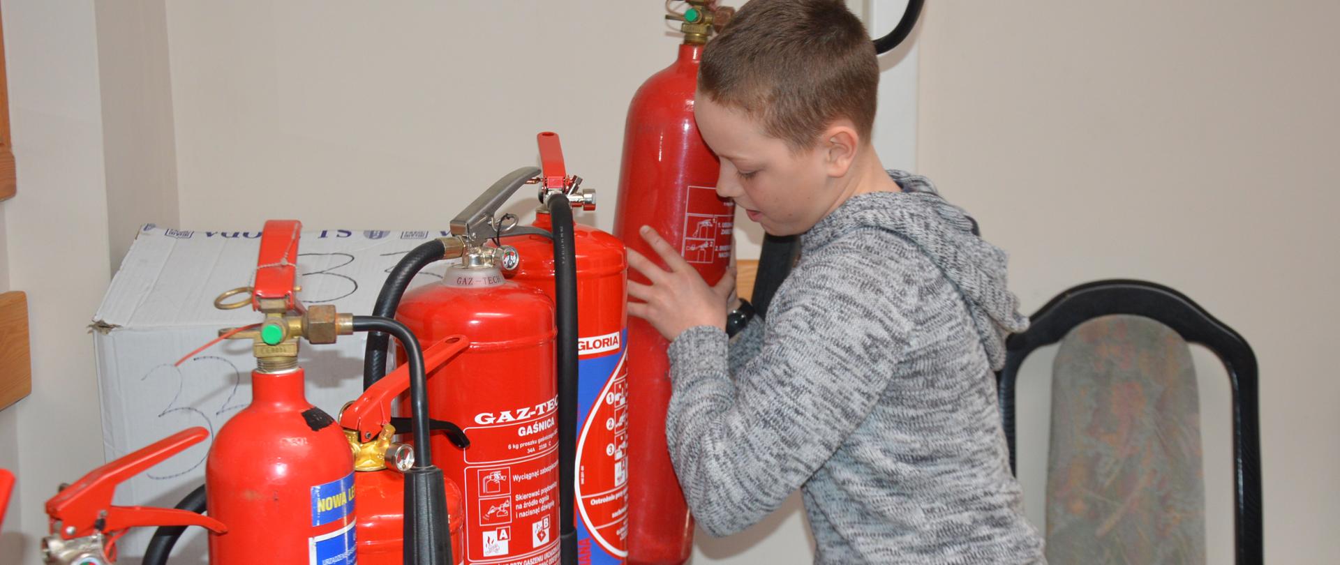 Eliminacje powiatowe OTWP. Garaż wozów bojowych. Uczestniczka eliminacji odpowiada na pytania dotyczące sprzętu ratowniczego. W rękach trzyma prądownicę. Obok niej inny uczestnik eliminacji oraz dwóch jurorów - strażaków z jednostki ratowniczo-gaśniczej. W tle widoczny jest fragment wozu strażackiego oraz regał z ubraniami specjalnymi i hełmami.