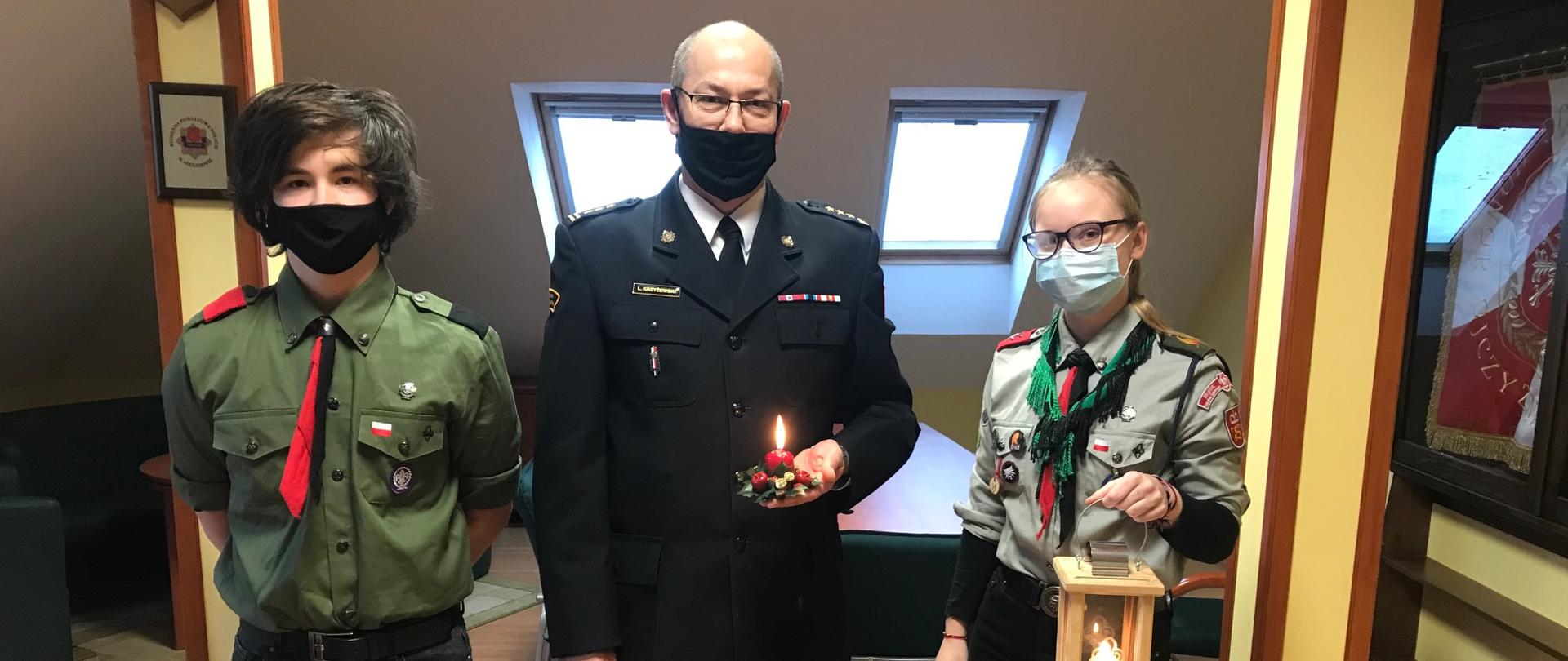 Na zdjęciu widać Komendata Powiatowego PSP w Augustowie oraz 2 harcerzy. Na zdjeciu widać palącą się świeczkę.
