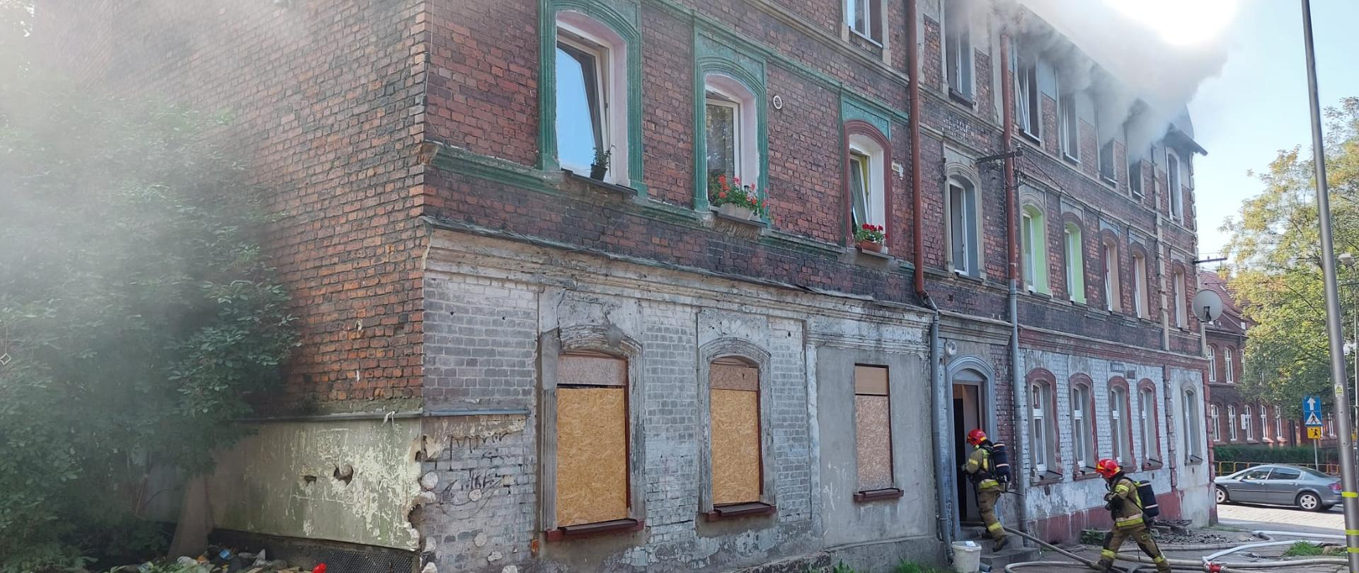 Dwóch strażaków w sprzęcie ochrony układu oddechowego wchodzi do środka budynku, z którego na drugim piętrze wydobywają się kłęby dymu.