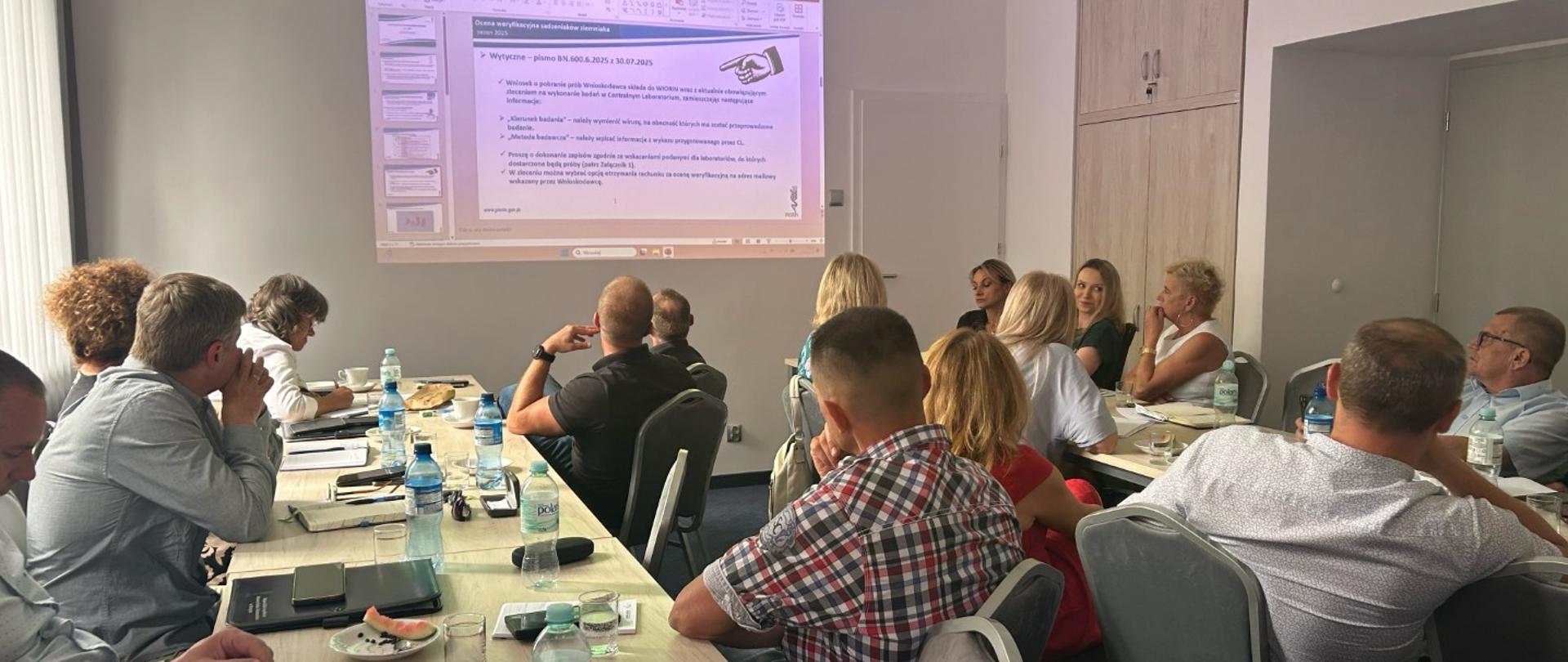 Zdjęcie przedstawia grupę osób siedzących przy stołach konferencyjnych w sali podczas spotkania lub szkolenia. Na ścianie wyświetlany jest slajd prezentacji multimedialnej.