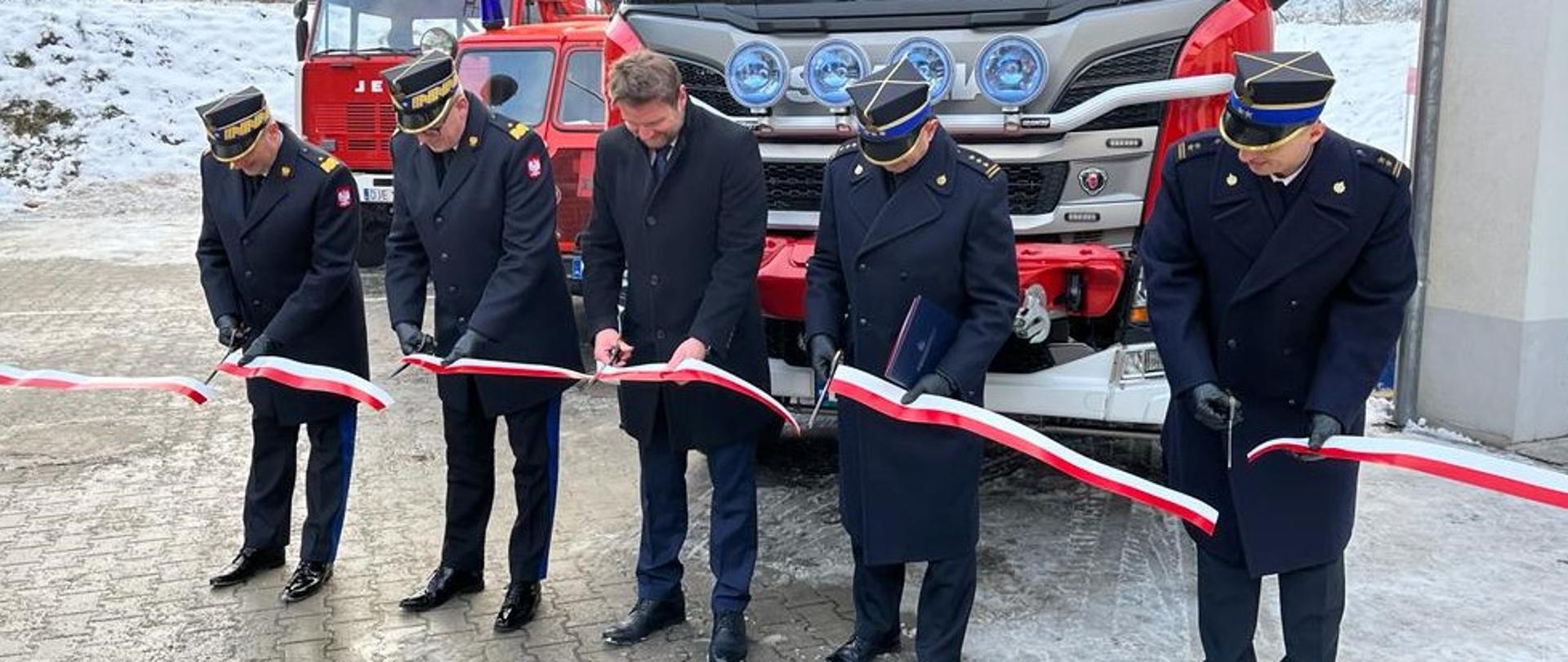 Na zdjęciu widać pięciu mężczyzn są to:
- nadbryg. Adam Konieczny- zastępca komendanta głównego Państwowej Straży Pożarnej,
- nadbryg. Krzysztof Hejduk- zastępca komendanta głównego Państwowej Straży Pożarnej,
- st. bryg. Marek Kamiński- dolnośląski komendant wojewódzki Państwowej Straży Pożarnej.
- bryg. Andrzej Ciosk - p.o. Komendanta Miejskiego Państwowej Straży Pożarnej w Jeleniej Górze.
- Radosław Jęcek - Burmistrz Miasta Karpacz.
Panowie przecinają wstęgę przed samochodem, który zostaje w ten dzień przekazany do służby. 