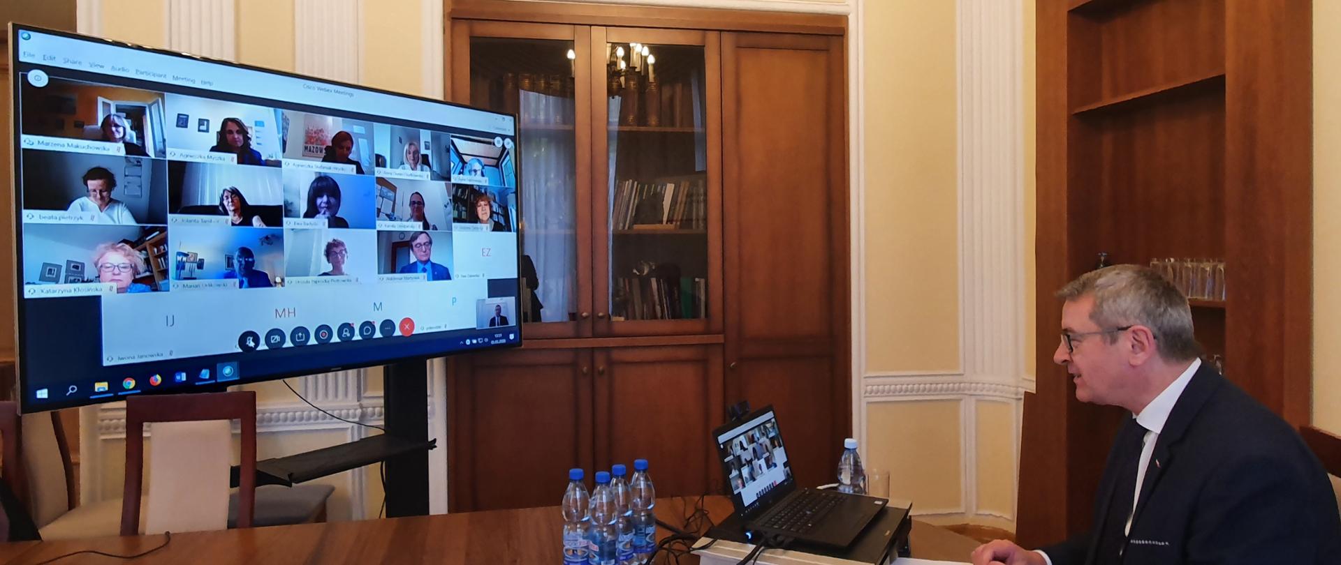 Minister Murdzek podczas telekonferencji, rozmawia z widocznymi na dużym monitorze członkami komisji.