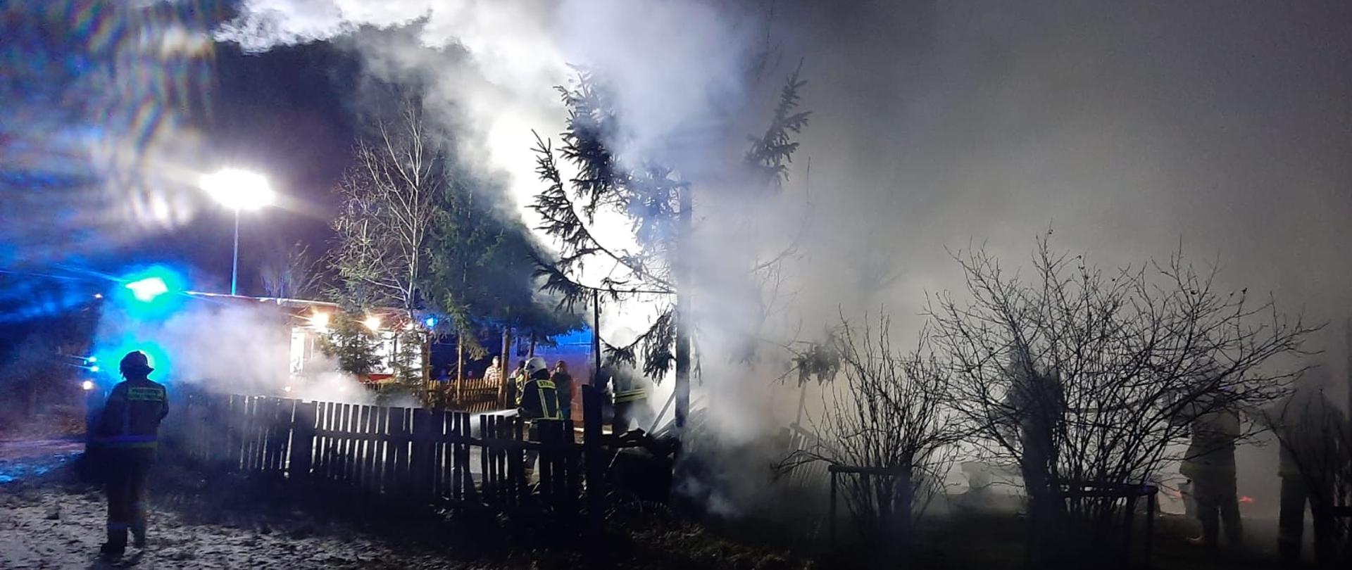 Palący się budynek gospodarczy. Wokół budynku strażacy oraz rozwinięte linie gaśnicze. Teren działań oświetla najaśnica. W tle samochód gaśniczy.