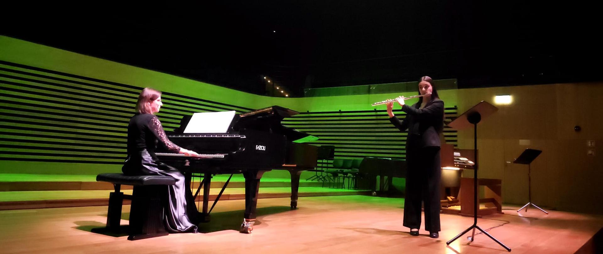 Nastolatka gra na flecie, akompaniuje jej na fortepianie kobieta.