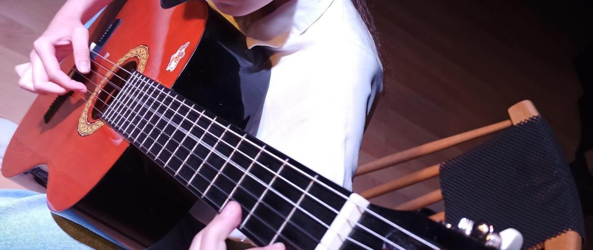 Zdjęcie przedstawia uczennicę siedzącą na krześle i grającą na gitarze zdjęcie jest wykonane od góry także na pierwszym planie widoczny jest gryf gitary w tle widać drewnianą podłogę
