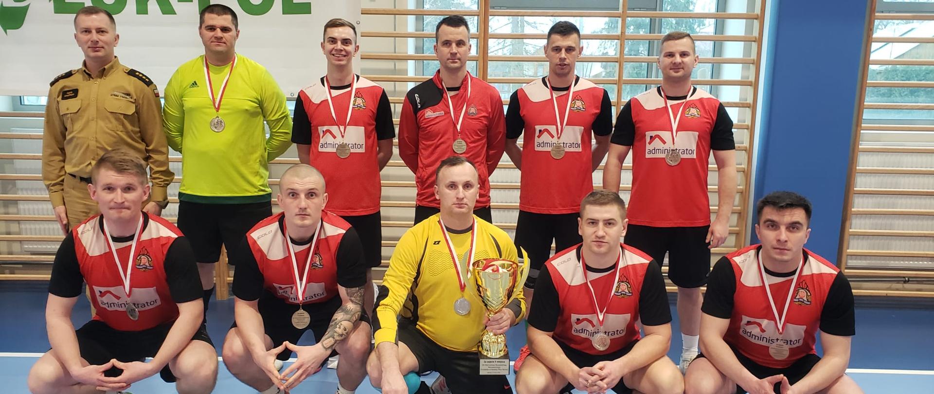 Zdjęcie wykonane na terenie obiektu sali sportowej w Węgrowie podczas rozgrywek finałowych strażaków w piłce nożnej. Na boisku widać ustawioną do pamiątkowego zdjęcia drużynę z komendy miejskiej Państwowej Straży Pożarnej w Radomiu, która zajęła drugie miejsce. Zawodnicy ubrani są w stroje sportowe w kolorze czerwono - czarno – białym z wyjątkiem dwóch bramkarzy którzy posiadają stroje w kolorach żółto – czarnym. 