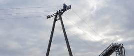 Na zdjęciu widoczny betonowy słup energetyczny linii średniego napięcia. Na jego szczycie siedzi mężczyzna. Obok znajduje się podnośnik hydrauliczny gotowy do podjęcia poszkodowanego. W koszu podnośnika dwóch strażaków w ubraniach specjalnych.