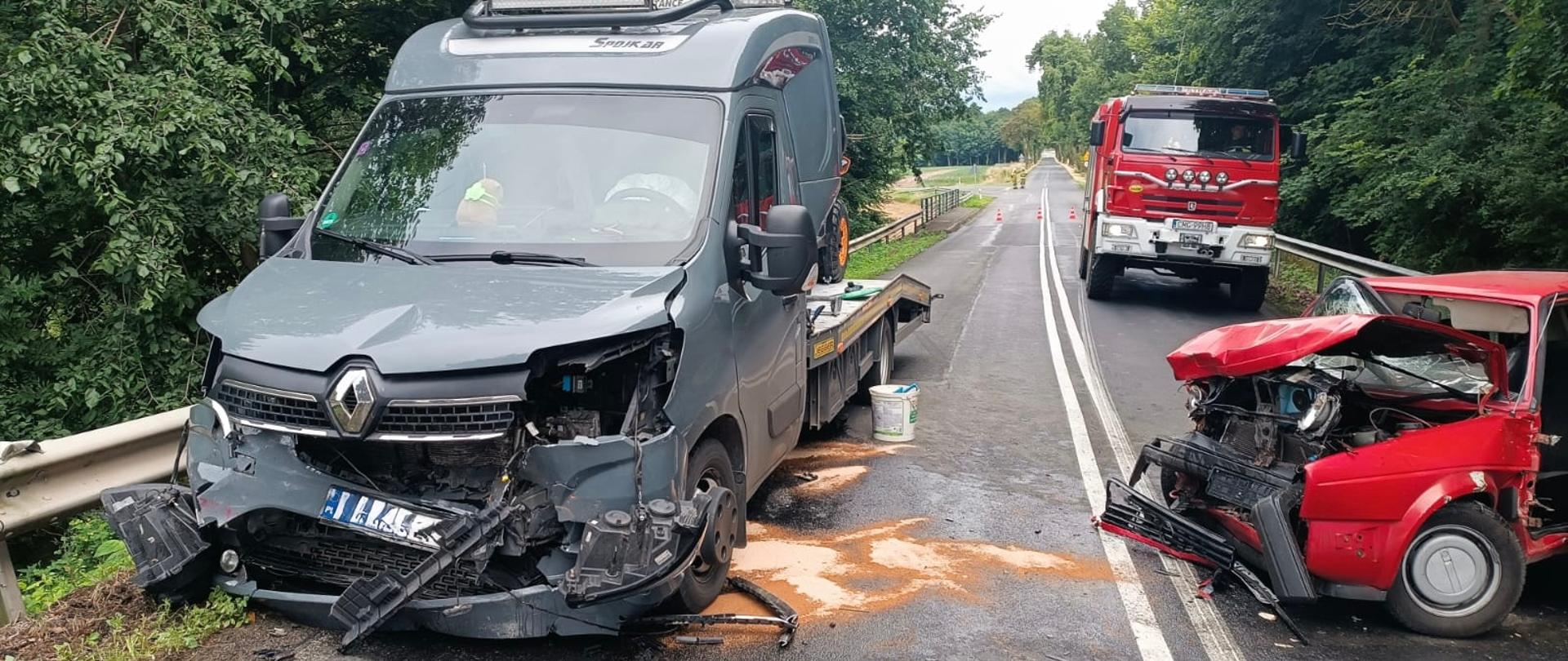 Na lewym pasie drogi stoi autolaweta. Na prawym pasie stoi samochód osobowy koloru czerwonego z rozbitym przodem. Za nim wóz strażacki.