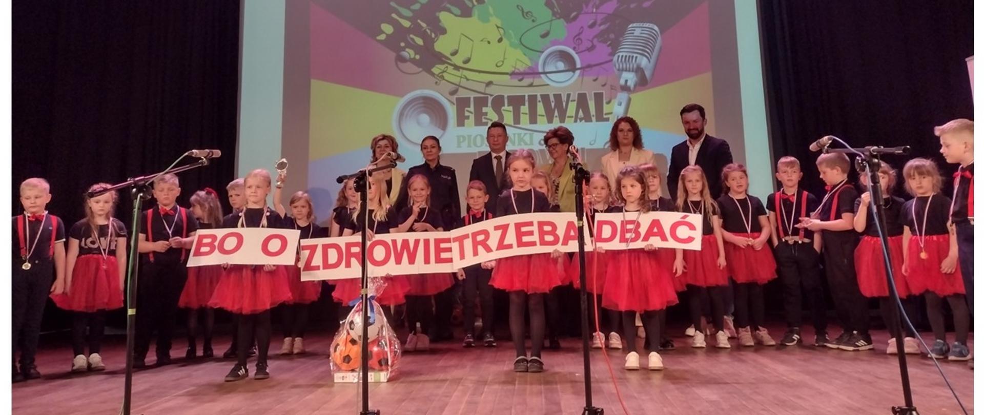 Zdjęcie z Festiwalu Piosenki o Zdrowiu 2026 organizowanego przez PSSE w Garwolinie. Dzieci stoją na scenie w kolorowych strojach, prezentują napis "bo o zdrowie trzeba dbać". Za dziećmi stoją Dorośli uczestnicy Festiwalu.