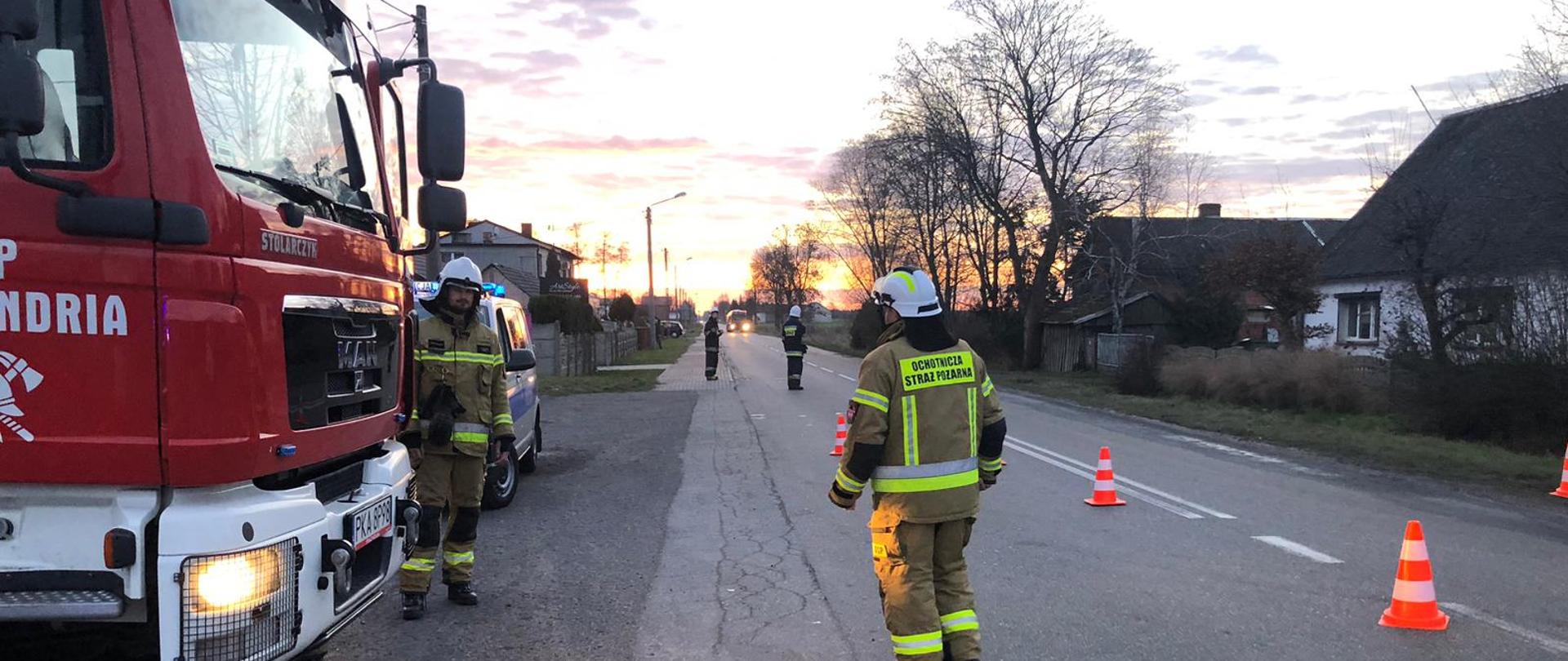Na zdjęciu droga publiczna. Widać umundurowanego funkcjonariusza. Na zdjęciu samochód ratowniczo-gaśniczy. Na drodze oznakowanie poziome. 