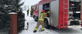 Strażak otwierający skrytkę w wozie strażackim. Wóz stoi na zaśnieżonej drodze. W oddali widać karetkę pogotowia i strażaków.