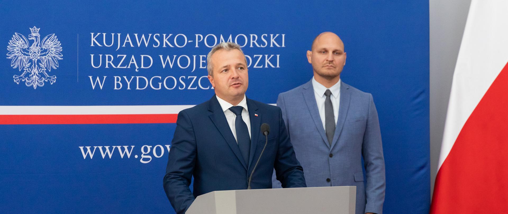 Jeden mężczyzna stojący przy mównicy i wypowiadający się, za nim drugi mężczyzna