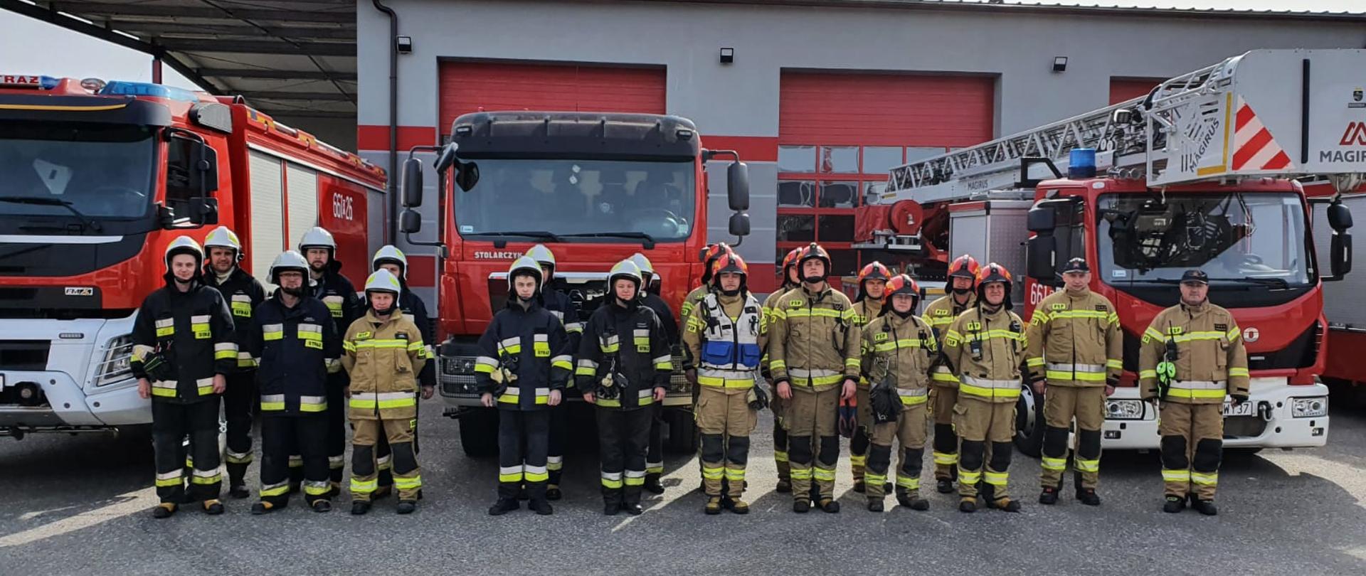 Na zdjęciu strażacy uczestniczący w ćwiczeniach na zbiórce przed ćwiczeniami w tle widoczne są samochody strażackie. 