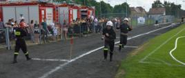 XI Powiatowe Zawody Sportowo-Pożarnicze OSP i MDP. Stadion w Miejskiej Górce. Na bieżni rozgrwana jest sztafeta pożarnicza 7x50 m z przeszkodami. Zawodnicy biegnący po torze wewnętrznym znajdują się w strefie zmian, tuż po przekazaniu prądownicy. Mają białe hełmy. Z lewej strony, na tle samochodów pożarniczych znajdują się kibice i strażacy. 
