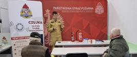 Zdjęcie przedstawia strażaka w jasnopiaskowym ubraniu specjalnym, który prowadzi prelekcję dla dwóch starszych mężczyzn siedzących w ławkach. Strażak stoi za podłużnym stołem, na którym umieszczone są dwie czerwone gaśnice oraz mniejsze materiały edukacyjne. W tle widoczna jest duża czerwona ścianka reklamowa Państwowej Straży Pożarnej, a po lewej stronie kadru stoi pionowy baner z dużym napisem „CZUJKA”.