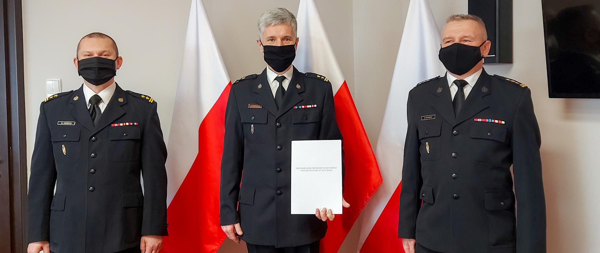 Na zdjęciu trzech strażaków, w tle trzy flagi Polski