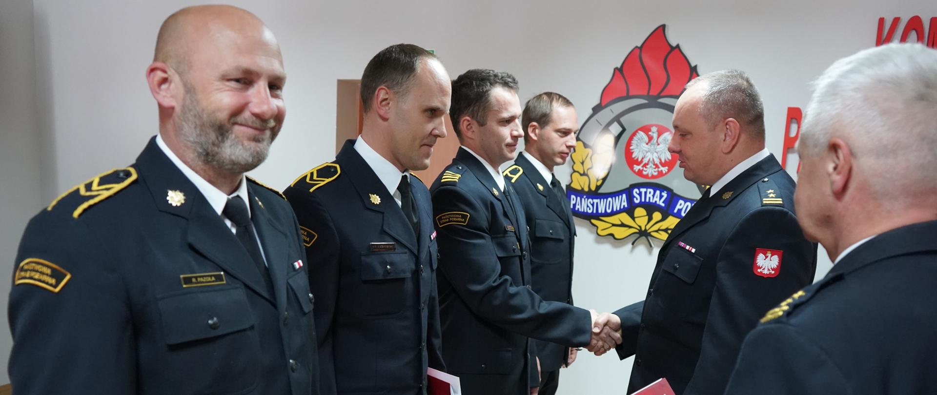 Świetlica Komendy Powiatowej PSP w Rawiczu. W szeregu stoi 4 awansowanych strażaków. Na przeciwko nich znajduje się komendant powiatowy wraz z zastępcą, którzy wręczają stosowne decyzje. Wszyscy mają na sobie umundurowanie wyjściowe. 