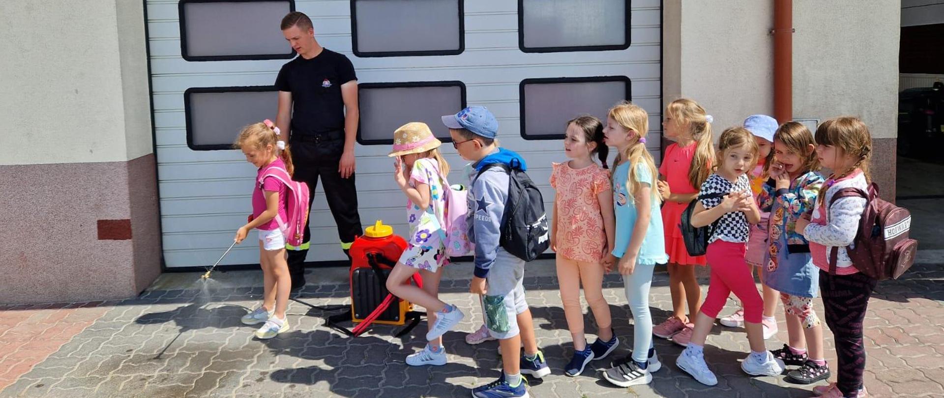 Grupa dzieci stojących na kostce brukowej, jedna leje wodą. W tle brama garażowa 