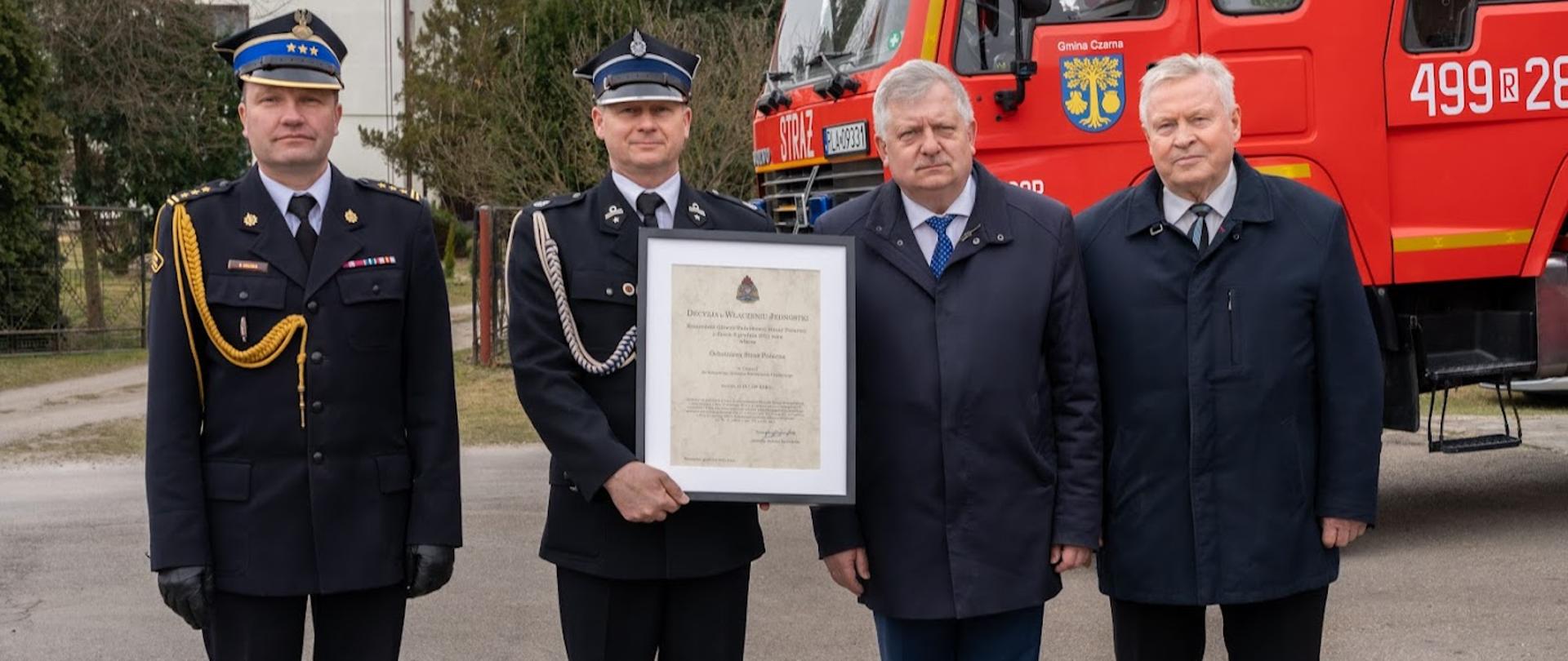Zdjęcie przedstawia prezesa Ochotniczej Straży Pożarnej druh Sławomir Chudzik w Czarnej trzymającego akt włączenia Ochotniczej Straży pożarnej w Czarnej. Obok stoją: poseł na Sejm Rzeczypospolitej Polskiej Kazimierz Gołojuch, komendant powiatowy Państwowej Straży Pożarnej w Łańcucie starszy brygadier Bogusław Golenia oraz wójt Gminy Czarna Edward Dobrzański. W tle ciężki samochód ratowniczo-gaśniczy Volvo. 