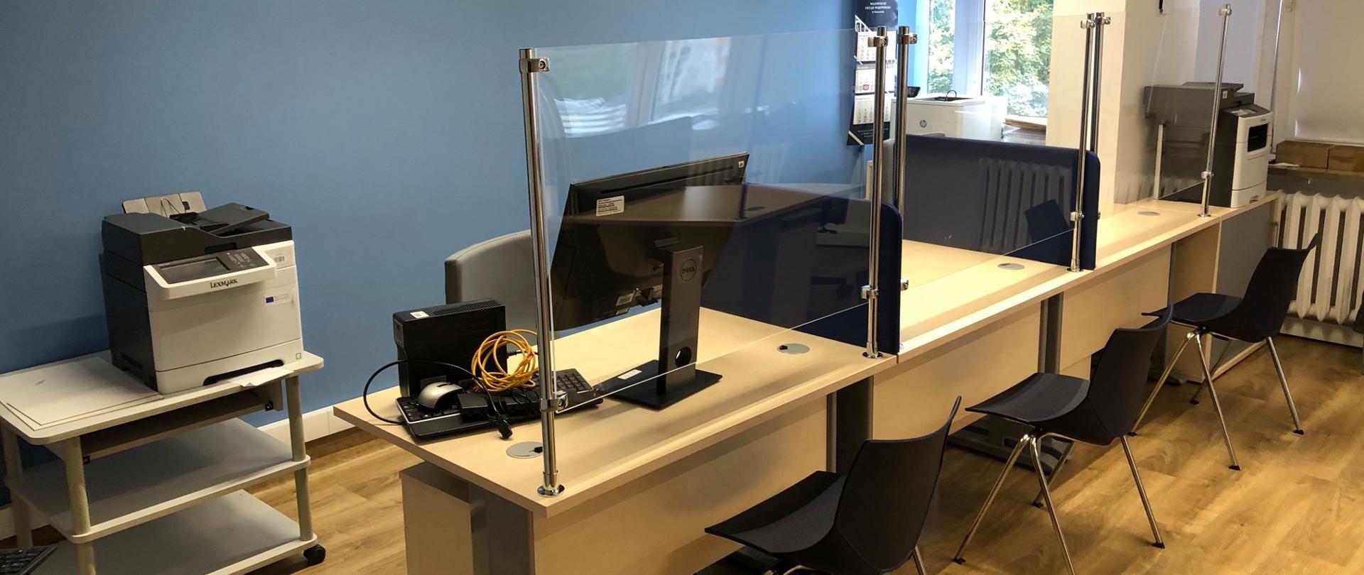 Pomieszczenia do obsługi klienta. Na środku widoczne trzy biurka, na których stoją komputery. Przy biurkach trzy krzesła dla klientów. Biurka od miejsca dla klientów oddzielone szybami. Po bokach widoczne stoliki z drukarkami. 