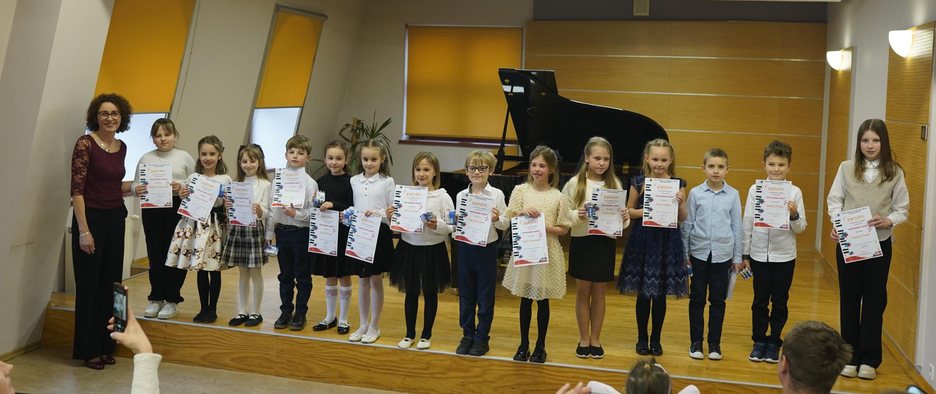 Wszyscy wykonawcy koncertu stoją w jednym rzędzie na scenie sali kameralnej, z lewej strony stoi nauczycielka prowadząca koncert.. Na pierwszym planie publiczność. Za uczniami fortepian oraz trzy okna przysłonięte roletami