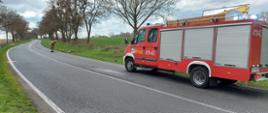 Na fotografii znajduje się wóz strażacki uczestniczący w działaniach, który stoi na jezdni po prawej stronie. Po lewej stronie strażak zamiata pozostałości płynów na jezdni. W tle znajdują sie drzewa i pole.